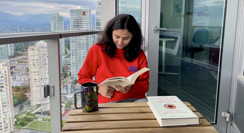 A woman is sitting at a table on a balcony reading a book. A mug and another book are sitting on the table.