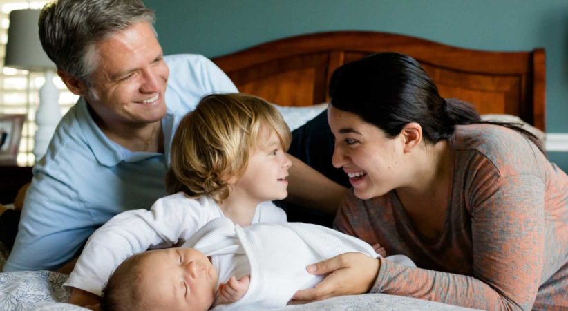 Two adults, one toddler and one baby smiling and lying on a bed together.