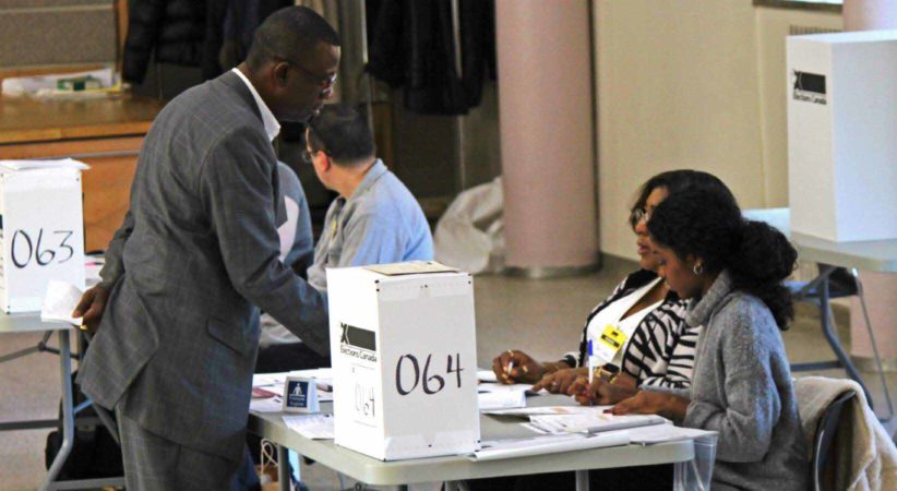 A man stands at a voting station with two women seated at the table.