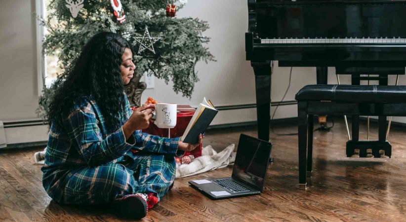 A woman reads a book while sitting in front of a Christmas tree and piano.