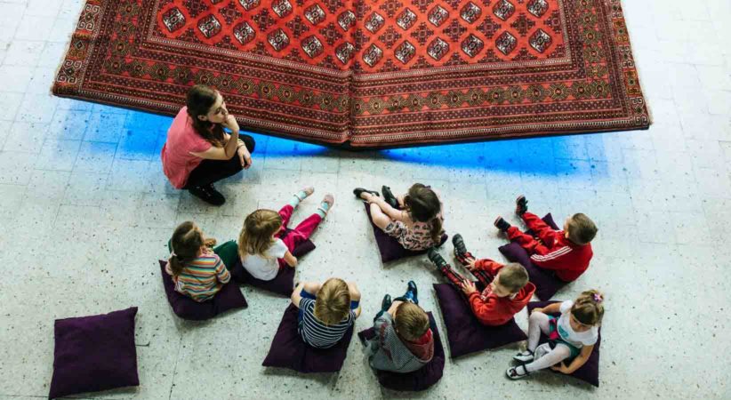 A group of children sit listening to an adult in front of an ornamental rug.