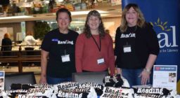 Three women stand smiling behind a table with t-shirts that say reading with a heart for the letter "I".