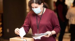 A woman wearing a medical mask drops a piece of paper into a box.
