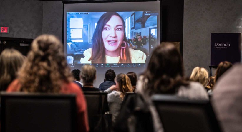 Angela Sterritt speaking via Zoom on a large screen in front of a crowd.