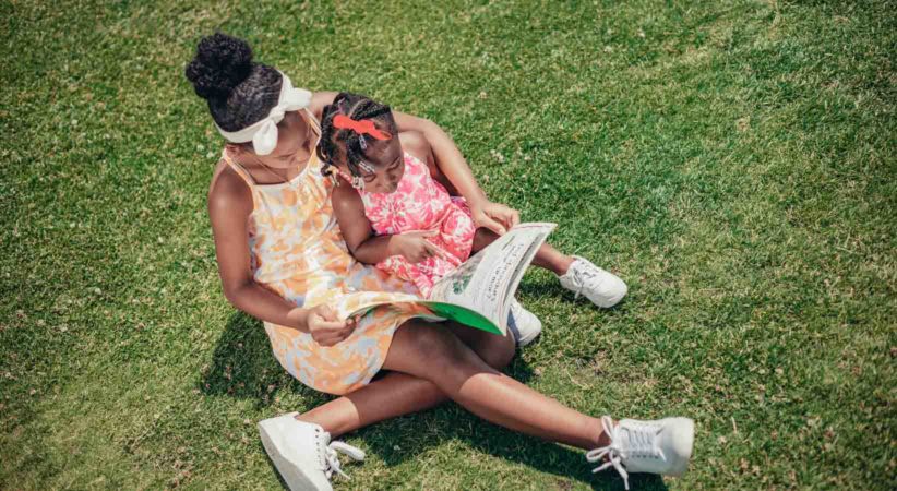 Two young girls sit on green grass reading a picture book together.