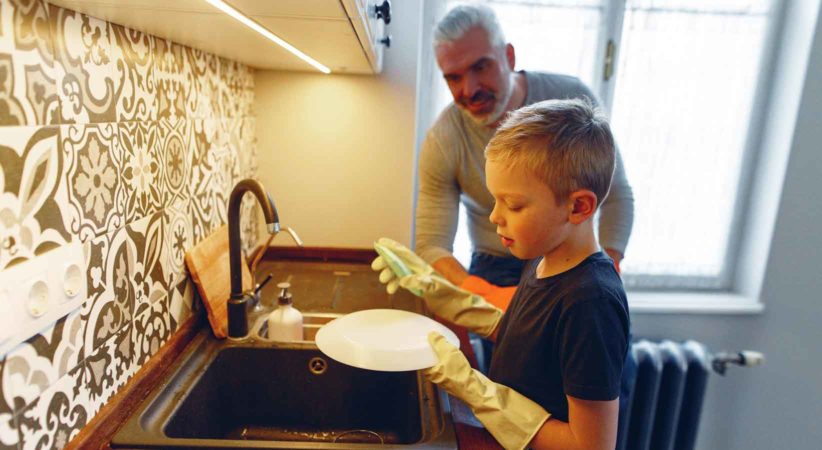 A boy washes dishes at a sink while a man stands behind him.