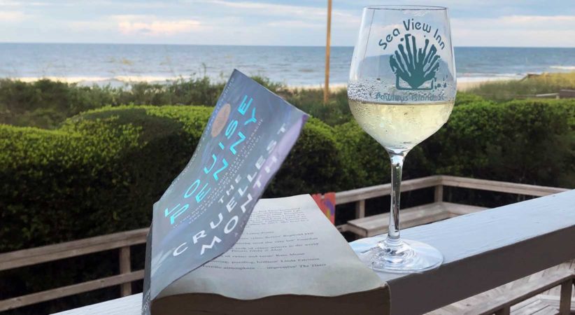 A book is lying on a balcony railing beside a glass of wine. A view of the ocean is in the background.