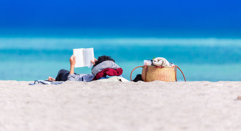 A person is lying on their back on a beach holding up a book. Vivid blue water can be seen in the background.