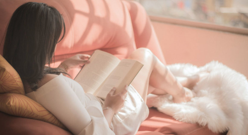 A woman lays on a pink couch reading a book. A blanket is at her feet.