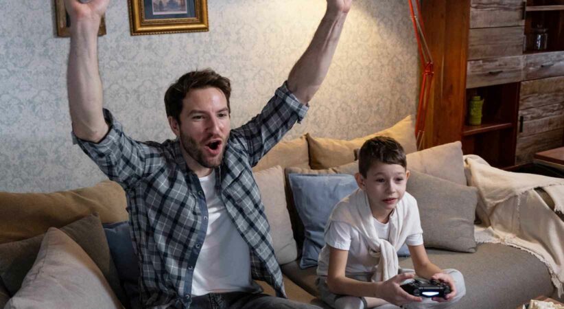 A man and a child playing video games while sitting on a couch.