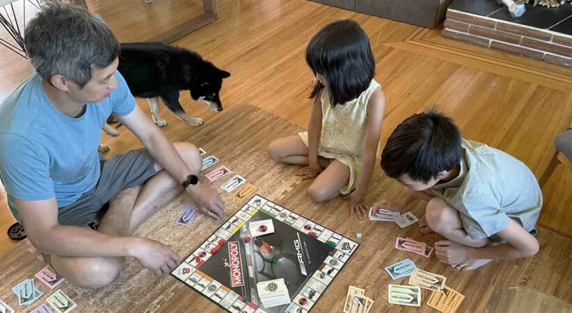 A man, dog, and two children are sitting on the floor playing the board game Monopoly.