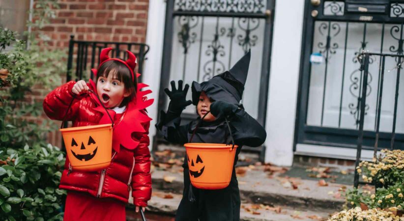Two children in costumes hold pumpkin-shaped buckets in front of a house