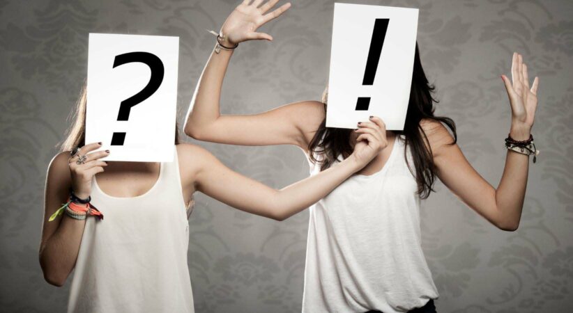 Two women holding paper in front of their faces. One has a question mark and the other has an exclamation mark.