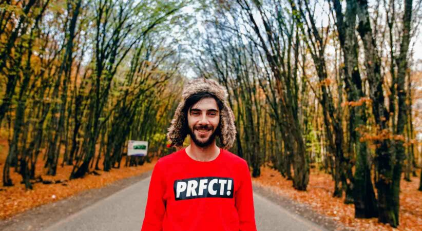 Man standing on a road with trees surround and wearing a red shirt with the word "perfect" missing the vowels.