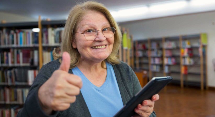 A senior woman holds an tablet in one hand and gives the camera a thumbs up with the other. A library with bookshelves is visible in the background.