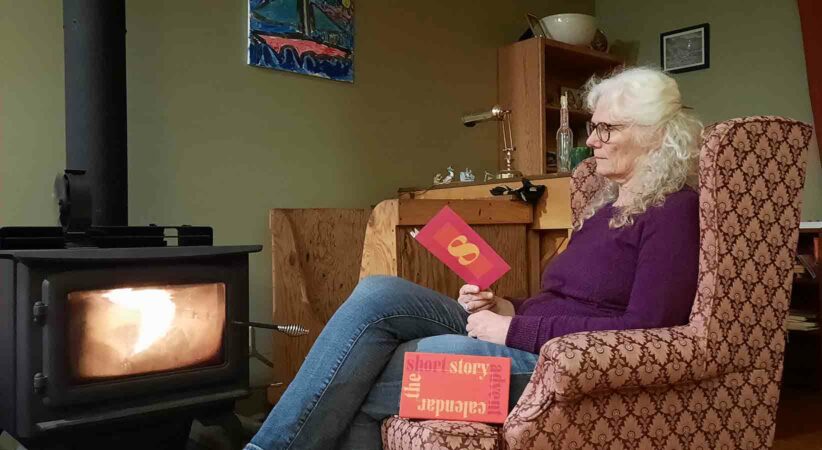 A profile of a woman sitting in a chair beside a fire and reading a book.