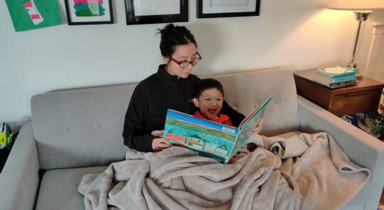A woman and child are reading a picture book on a couch under a blanket.