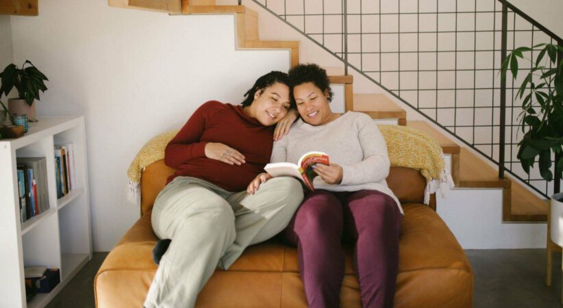 Two people, one pregnant, reading a book together on a couch