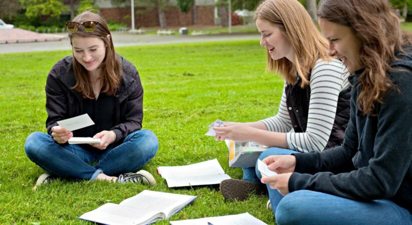 Three women are sitting cross-legged on grass while looking at flashcards.