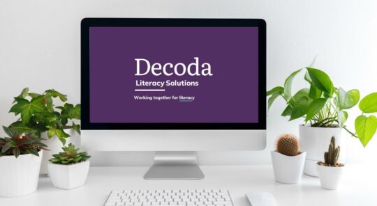 A desktop computer surrounded by plants and showing Decoda's logo and tagline on the screen.