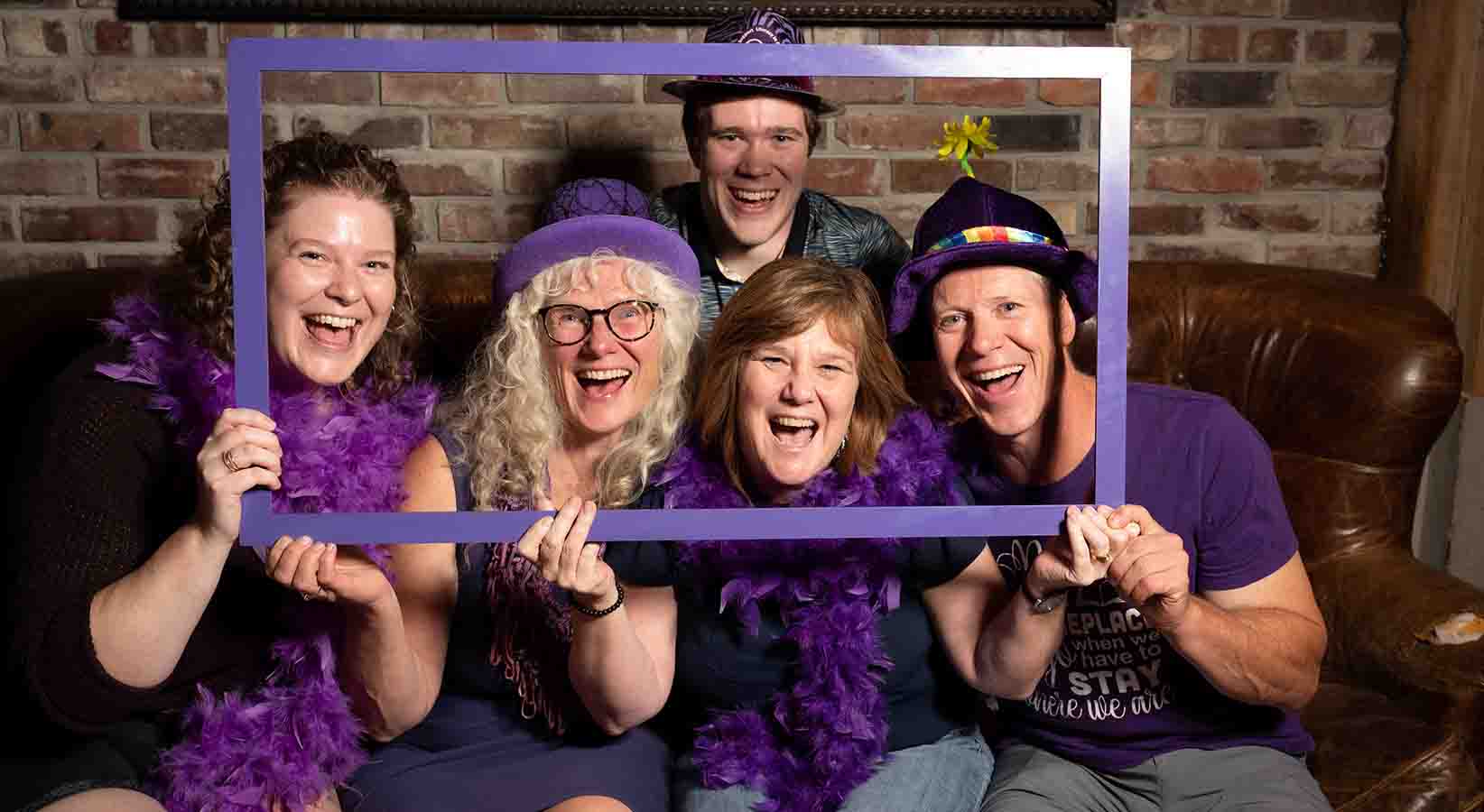 A group of people wearing purple props hold a purple frame around their faces while sitting on a couch.