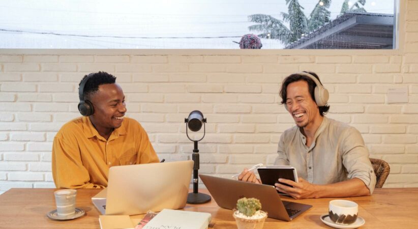 Two men are sitting at a table with podcasting equipment and smiling.