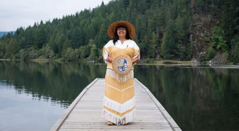 An Indigenous woman wearing traditional garb stands on a doc in front of a lake. She is holding a drum in front of her.