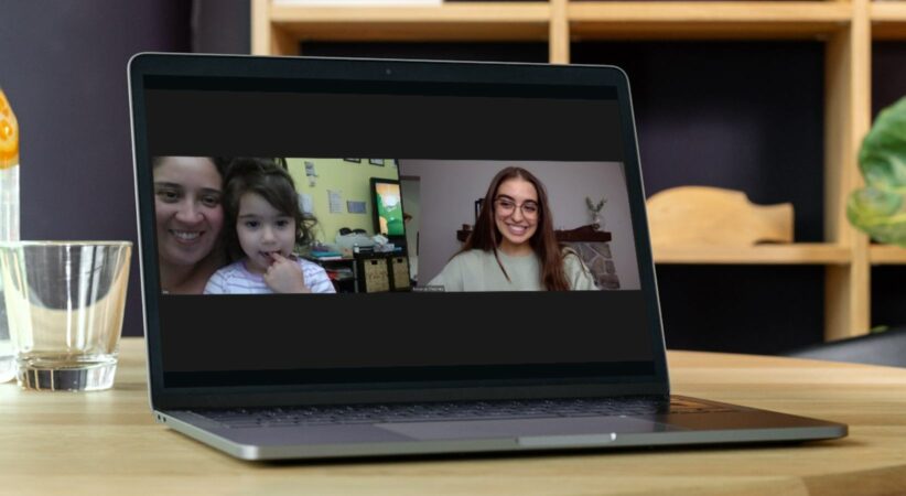 A laptop is sitting on a wooden desk. On the screen a woman and child, and another young woman, are on a video call.