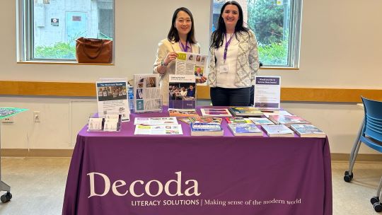 Two people are displaying materials at a table, the tablecloth says Decoda Literacy Solutions
