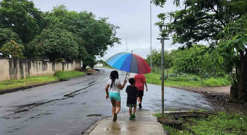 two children and an adult walking down a street while holding a rainbow-coloured umbrella.