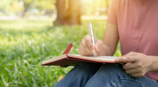 A person sitting in grass holding a pen and notebook.