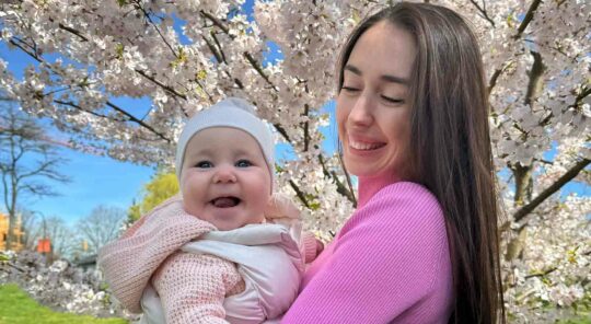 A woman in a pink shirt holds a smiling baby in front of a cherry tree in bloom