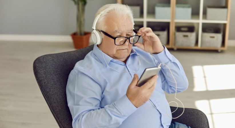 A senior man is sitting holding a device and wearing headphones