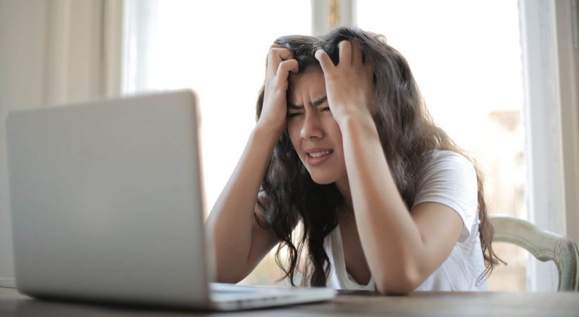 Frustrated women with her hands clasping her hair, grimaces at a laptop screen.