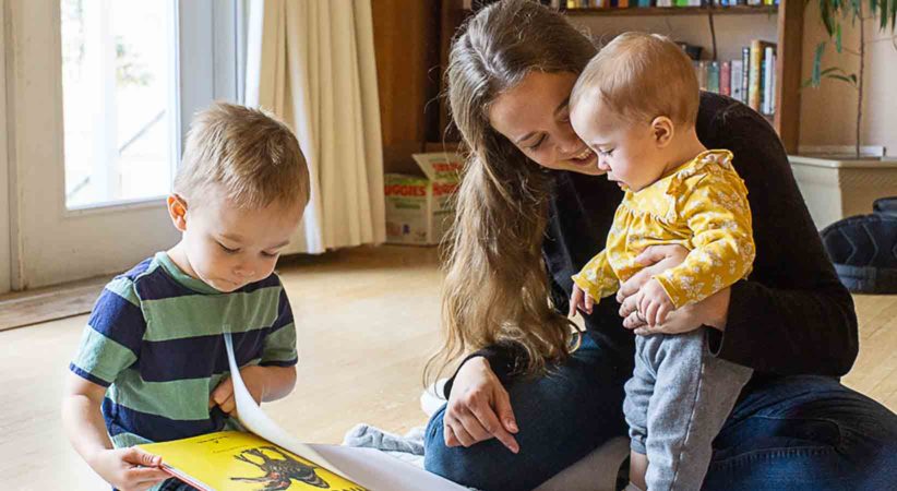 A mother holds a baby while a toddler reads a book