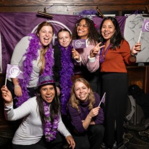 Six women - four standing and two crouching, dressed in purple accessories